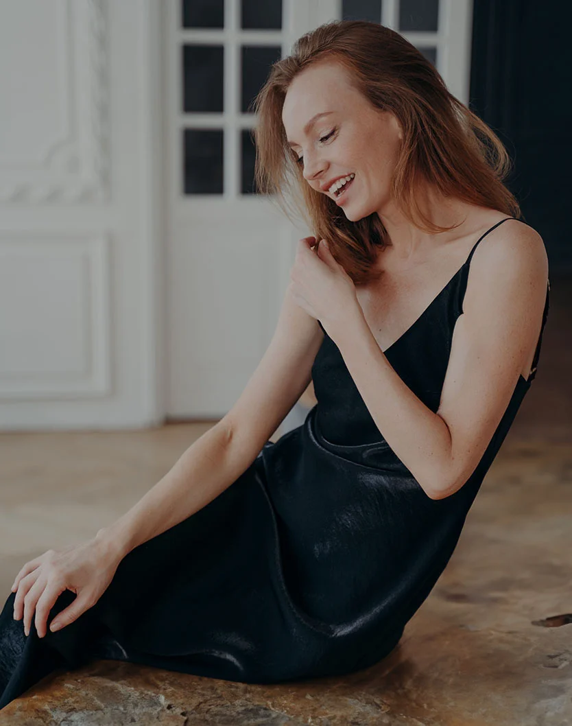 A light-skinned woman with shoulder-length, light brown hair sits on a light brown wooden surface, wearing a black, sleeveless midi slip dress. She smiles softly, looking slightly downward. The background includes pale white walls, light tan floor, and a window. Soft, diffused lighting creates an elegant, calm atmosphere. - Arm Lift in Beverly Hills, CA