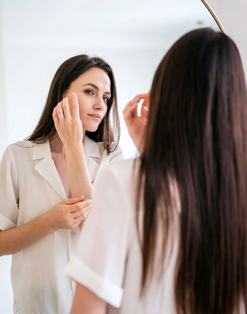 A woman with long, dark brown hair looks at herself in a mirror, wearing a loose-fitting, cream-colored short-sleeved top. She is touching her face, possibly applying makeup or skincare. The background features a light beige or white wall, with a rounded mirror reflecting her image. The focus is on her face and hands. - Browlift in Beverly Hills, CA