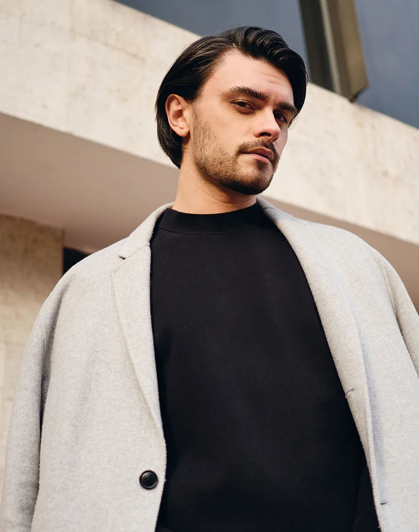 A man with short, dark brown hair and a light-medium complexion is shown from the mid-chest up. He has a short, trimmed beard and mustache, with a neutral to slightly serious expression. He wears a light gray, textured wool-like coat over a black crew-neck top. The background features a light beige/tan wall with horizontal lines, hinting at architectural design. - Earlobe Reduction in Beverly Hills, CA