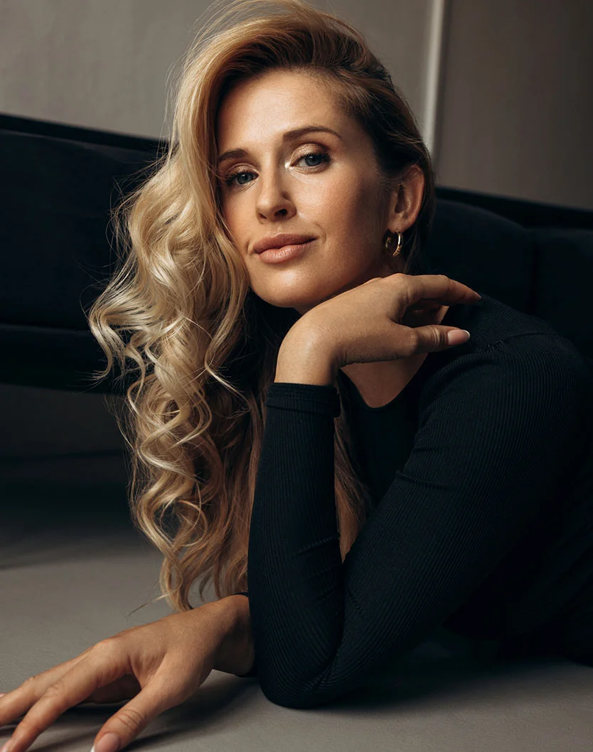 A woman with long, wavy blonde hair lies on a light gray surface, wearing a form-fitting black long-sleeve top. She gazes softly at the camera with a serene expression. One hand rests on her cheek, and the other extends in front of her. Soft lighting highlights her features and hair, with gold hoop earrings visible. The muted gray background keeps the focus on her. - Eyelid Surgery in Beverly Hills, CA