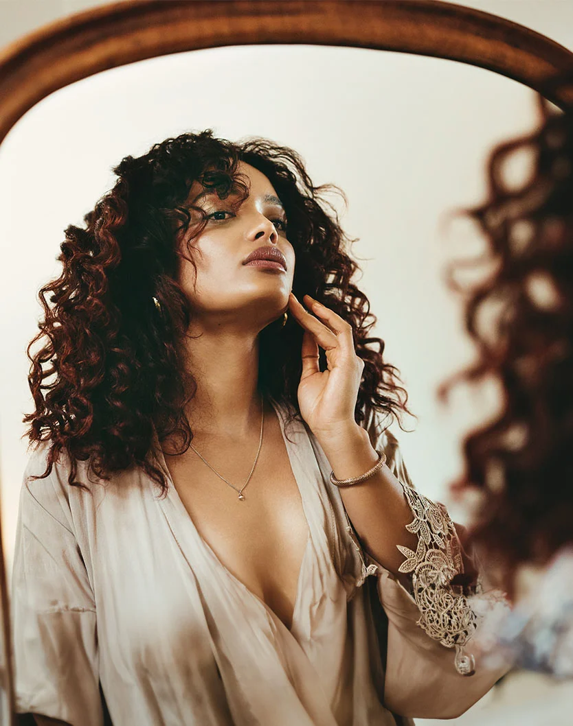 A woman with curly, dark brown hair looks at herself in a mirror. She wears a light beige, loose-fitting V-neck top with lace-trimmed sleeves and a delicate necklace. Touching her cheek with one hand, her expression is thoughtful and slightly upward. The mirror has a large, arched, light-brown wooden frame, and the soft, warm lighting creates a serene atmosphere. - Mole Cyst Keloid Removal in Beverly Hills, CA