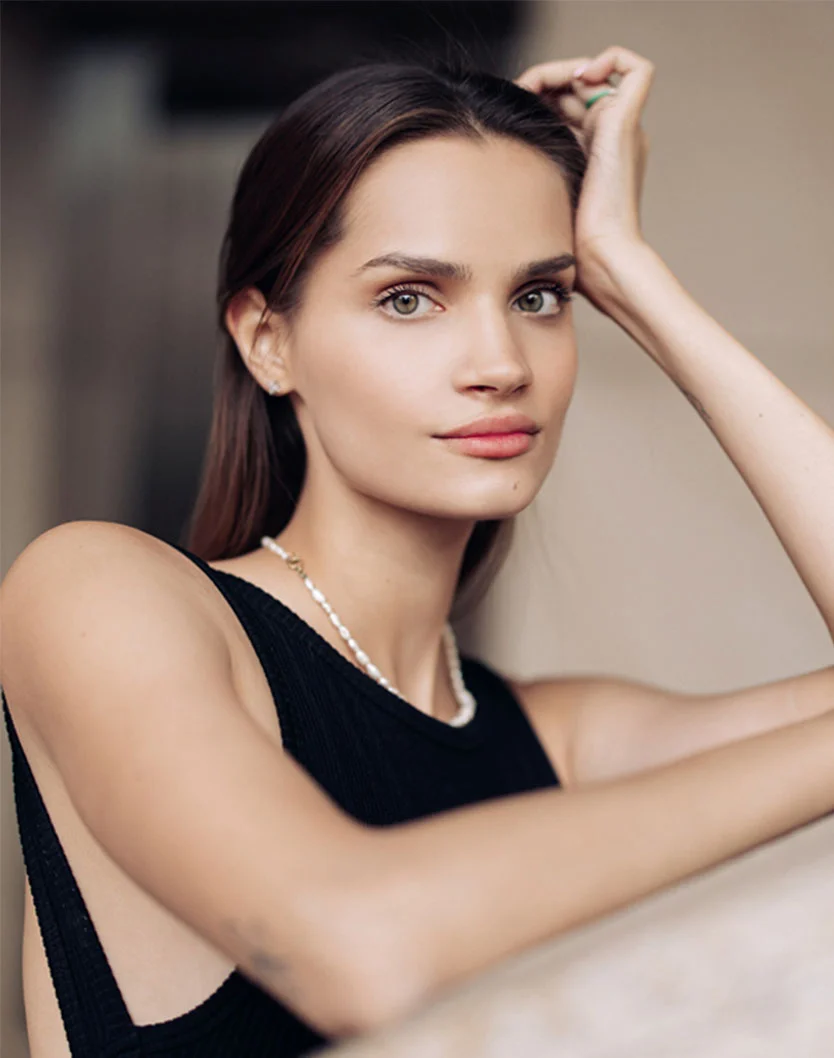 A woman with long dark brown hair and light-colored eyes poses against a soft beige background. She has a light complexion and a neutral, direct expression, looking slightly toward the viewer. Leaning slightly forward, she rests one hand on her head and the other on a surface. She wears a simple black sleeveless top, a delicate pearl necklace, and small stud earrings. - Otoplasty in Beverly Hills, CA